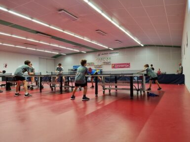 Jeunes joueurs pratiquant le tennis de table dans une salle adaptée. Trois tables alignées avec les joueurs.