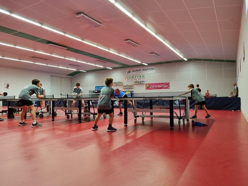 Jeunes joueurs pratiquant le tennis de table dans une salle adaptée. Trois tables alignées avec les joueurs.