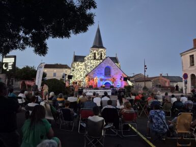 Spectateurs assis devant la scène positionnée sur le fronton de l'église de Marigny illuminée avec un jeu de lumière.