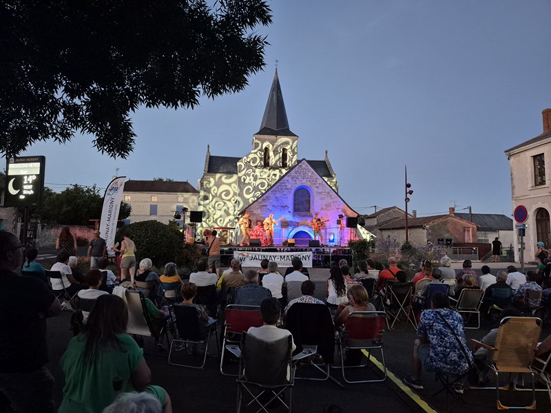 Spectateurs assis devant la scène positionnée sur le fronton de l'église de Marigny illuminée avec un jeu de lumière.