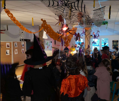 Enfants déguisés en sorcière, fantôme etc... dans une salle décorée avec des guirlandes oranges, toiles d'araignées et fausses araignées suspendus au plafond.