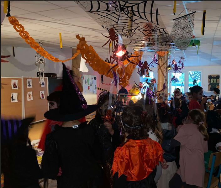 Enfants déguisés en sorcière, fantôme etc... dans une salle décorée avec des guirlandes oranges, toiles d'araignées et fausses araignées suspendus au plafond.