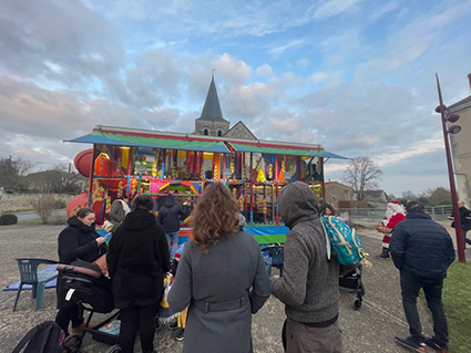 Personnes devant un manège devant l'église de Marigny-Brizay. En arrière plan, le Père Noël distribue des bonbons avec son panier à la main.