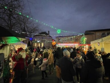 Place de la fontaine animée de plusieurs stands, décorations et sapin de Noël et manège avec au centre un public nombreux.
