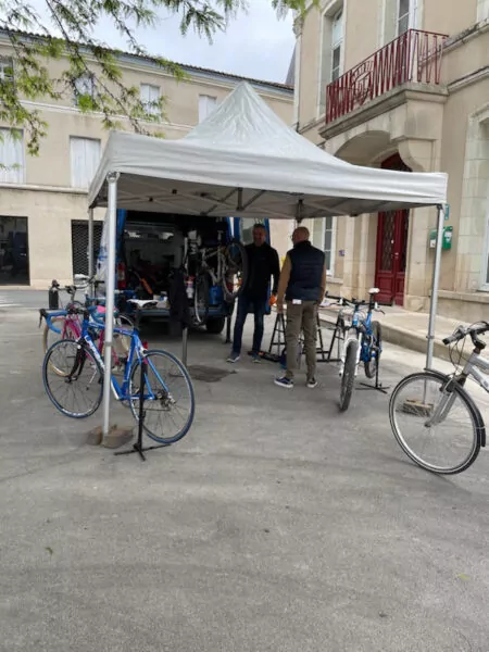 Stand positionné sur la place de la fontaine abritant plusieurs vélo en attente d'être révisés ou réparés. Deux personnes sont en train de regarder à un vélo monté sur un pied.