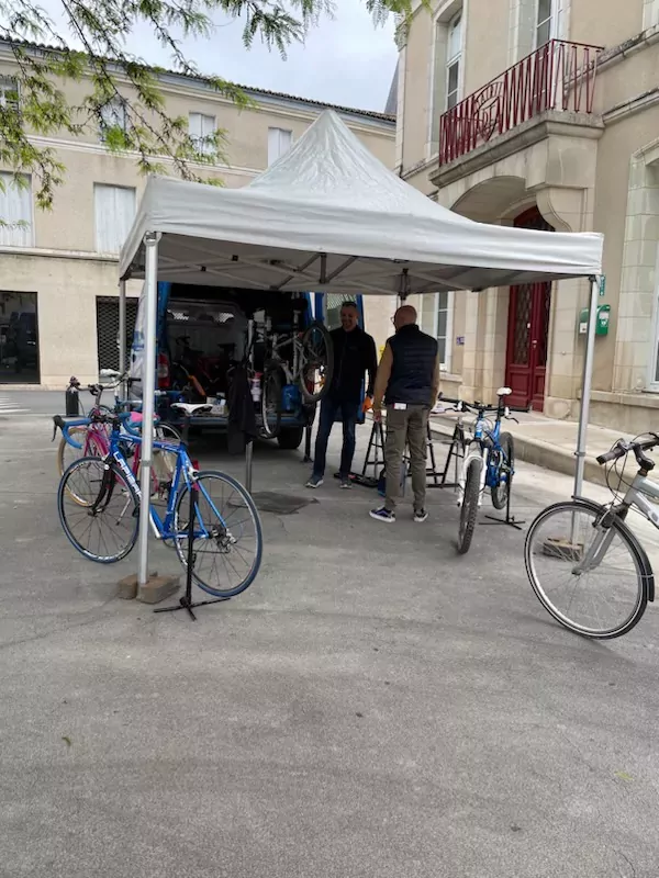 Stand positionné sur la place de la fontaine abritant plusieurs vélo en attente d'être révisés ou réparés. Deux personnes sont en train de regarder à un vélo monté sur un pied.