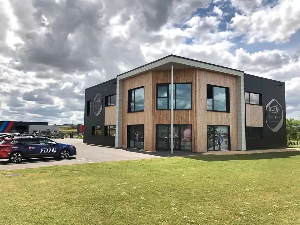 Bâtiment en bois abritant le siège de l'équipe cycliste internationale FDJ Suez sur le site d'activités de la Grand'Raise, au pied duquel est stationné un véhicule aux couleurs de l'équipe.