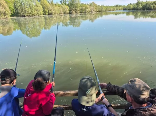 3 enfants vus de dos sur un ponton de pêche tiennent une canne. Ils sont accompagnés d'un adulte qui les guide.