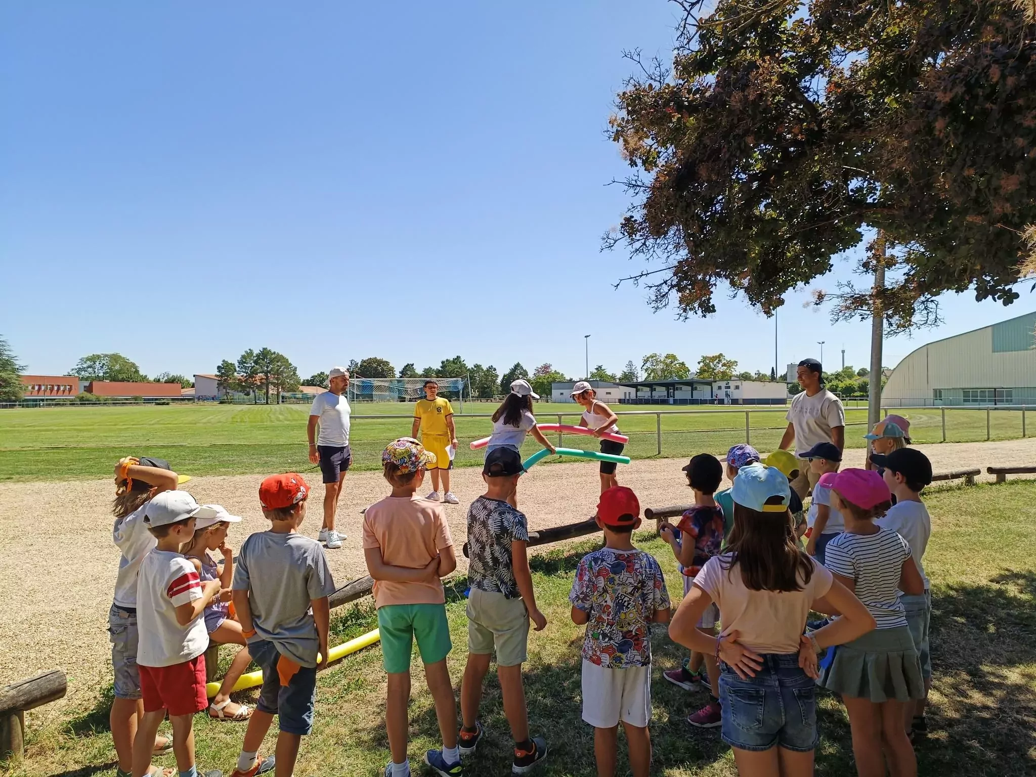 enfants qui se tiennent debout à l'ombre chacun avec sa casquette sur la tête regardant deux autres enfants s'affronter lors d'un jeu sur le complexe sportif.