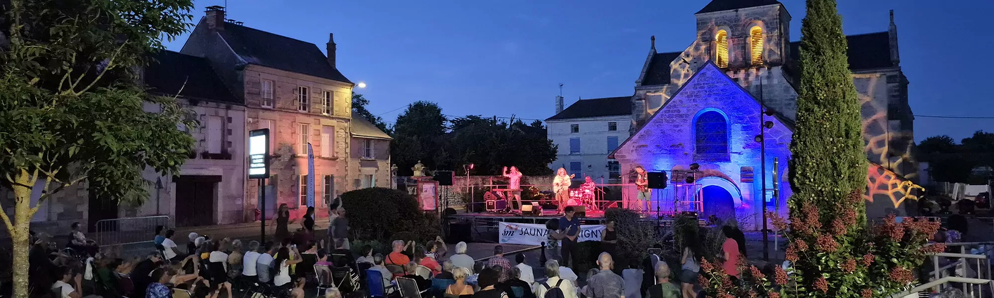 Concert à la tombée de la nuit devant l'église de Marigny-Brizay illuminée. Spectateurs assis devant la scène qui est elle même devant l'église.