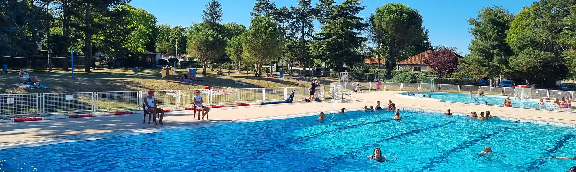 Vue sur la piscine, ses 2 bassins et son parc. Les maîtres-nageurs sont au bord du bassin et surveillent les baigneurs