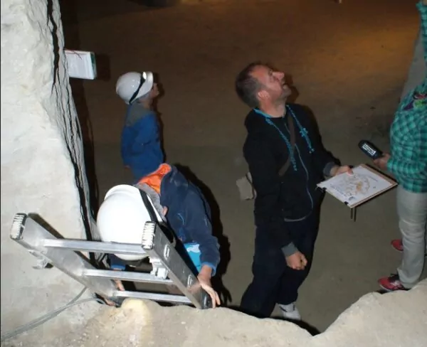 Deux enfants portant un casque de chantier avec une frontale, accompagnés d'un adulte, réalisent un jeu de piste dans une cave souterraine.